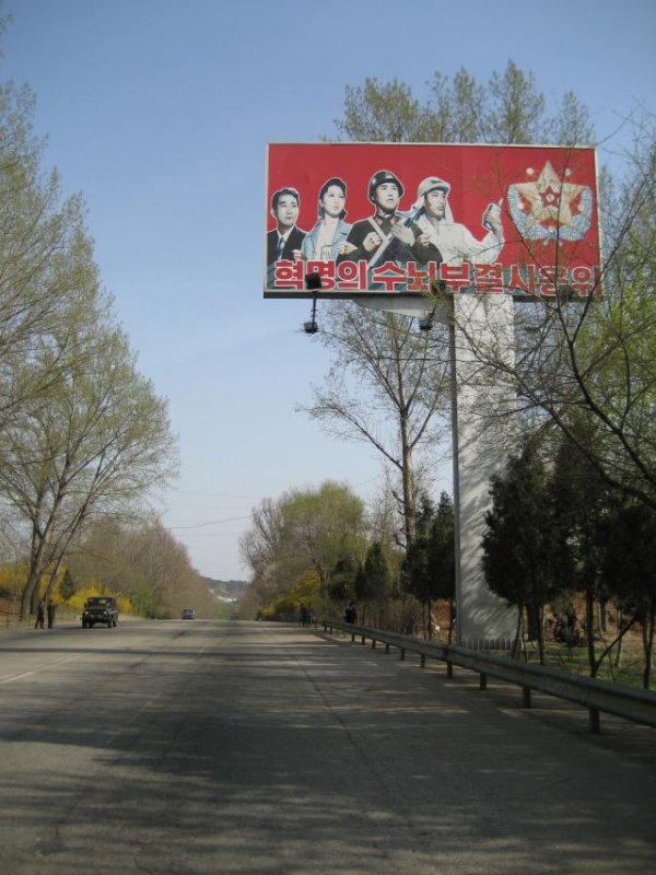 Pohjois-Koreassa ei ole mainoksia, mutta propagandajulisteita ja iskulauseita on pystytetty joka nimeen, notkoon ja saarelmaan. Valokuvassa harvinainen tilanne: kaksi autoa tiellä yhtä aikaa.