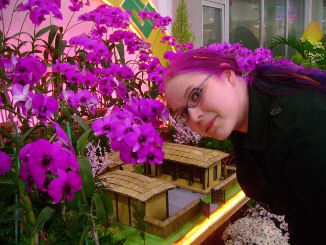 Toimittaja kukkanäyttelyssä. Kukka-asetelman keskellä pienoismalli Suuren Johtajan lapsuudenkodista.