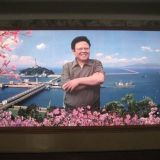 Kuva Kim Jong-Ilista vastavalmistuneen padon edessä. Valokuvissa Kimin hampaat ovat huonommassa kunnossa, rillit tyhmemmät ja pötsi isompi kuin tässä maalauksessa.